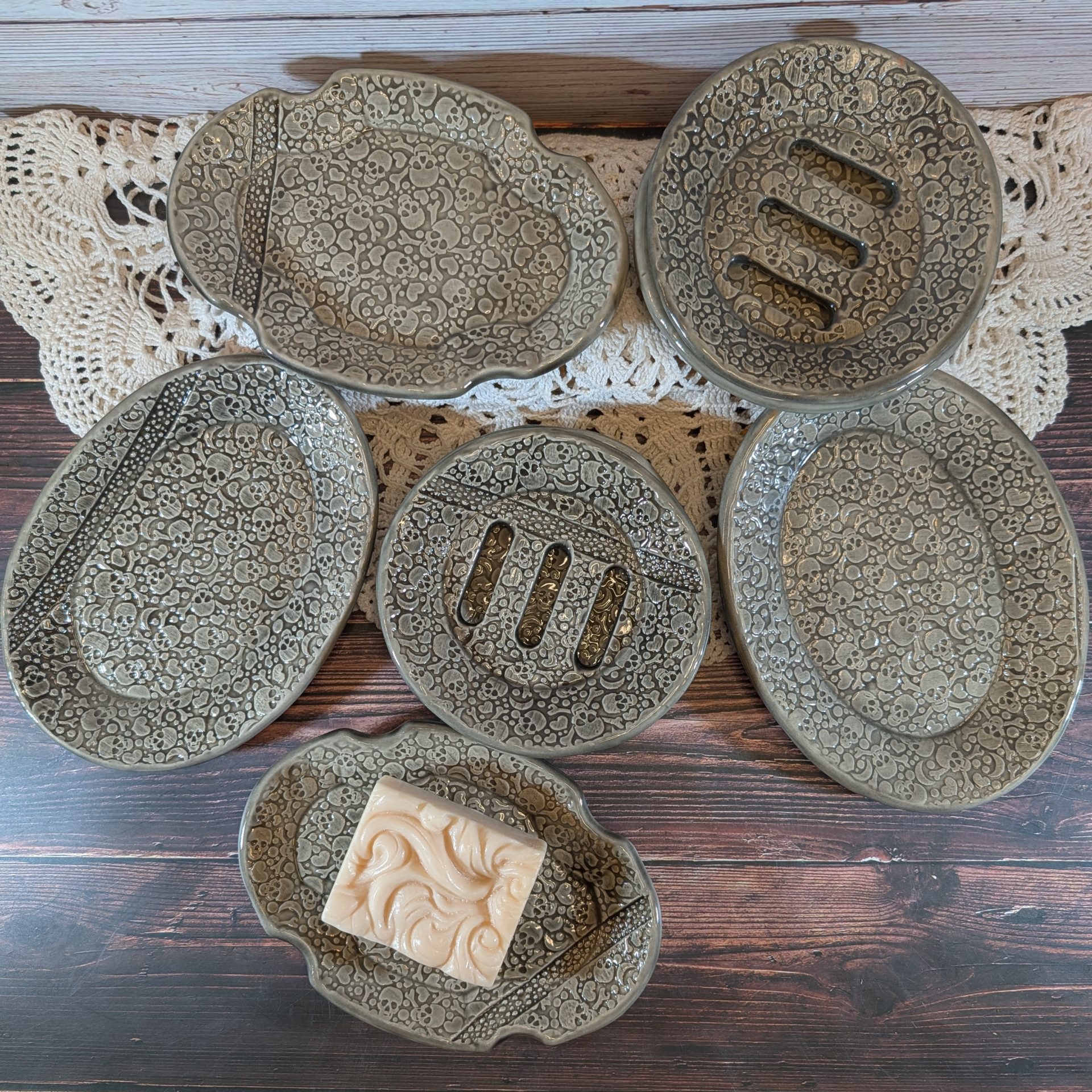 A selection of gray soap dishes, one with a bar of soap, on a wooden surface with a white lace doily.