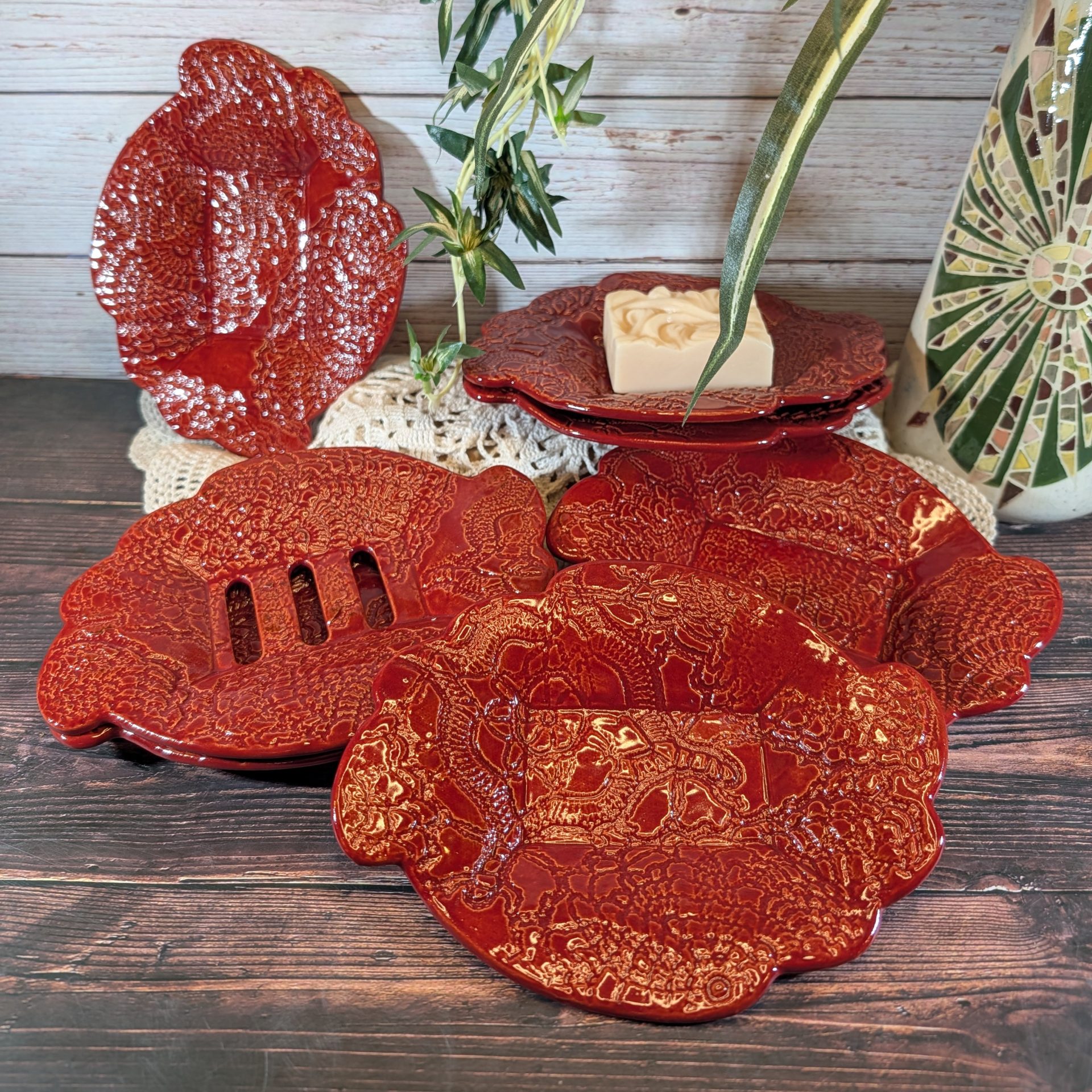 A selection of red soap dishes with a doily motif. They are on a white doily on a wooden surface, and one of them has a bar of soap in it.
