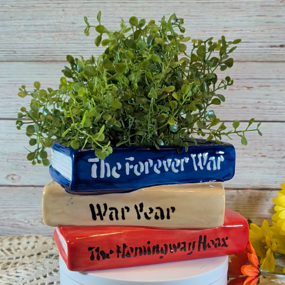 A ceramic planter that looks like a stack of three books, sitting on a white stand, with a wooden background behind it.