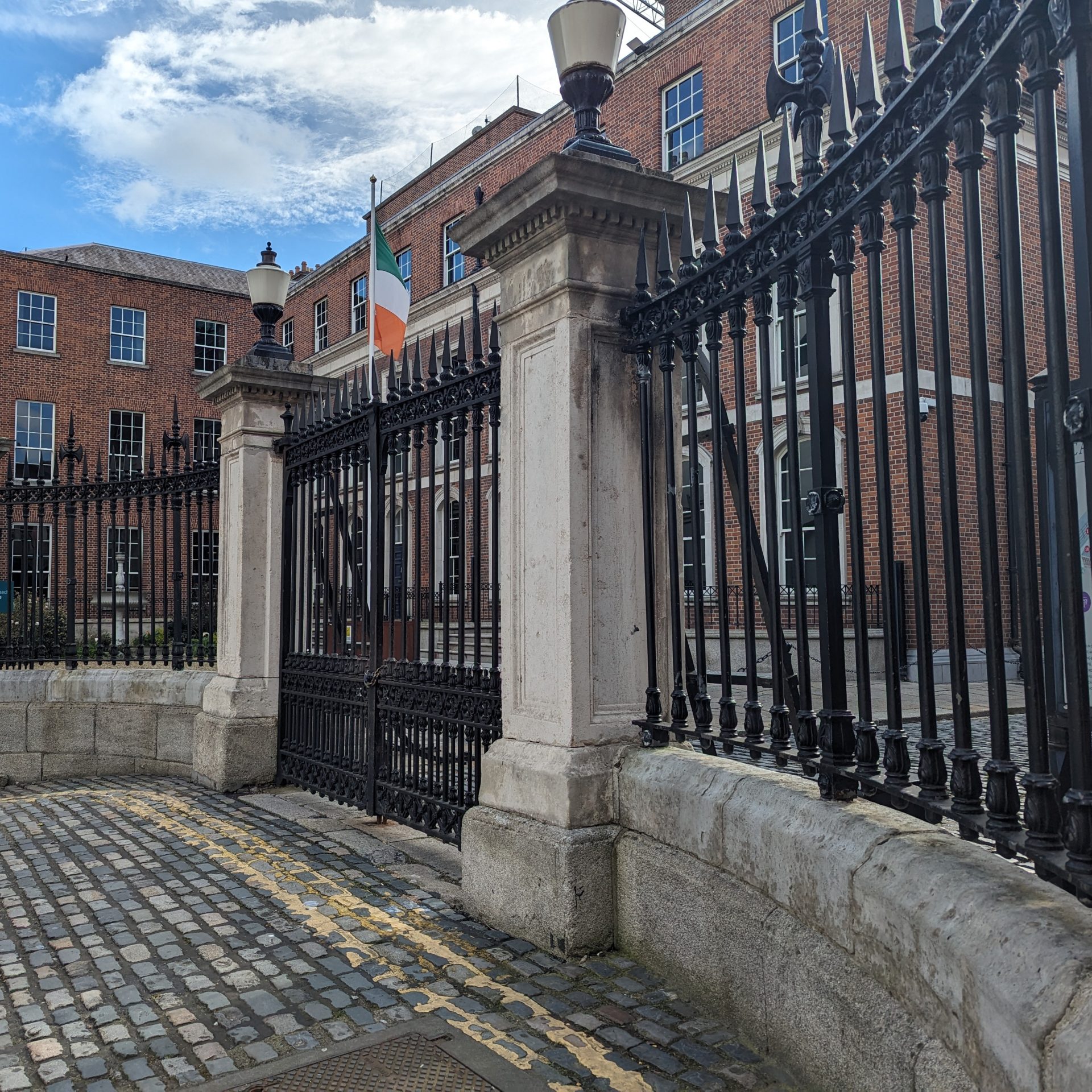 A curved stone and iron fence and gate on a cobblestone road, with a brick building behind.